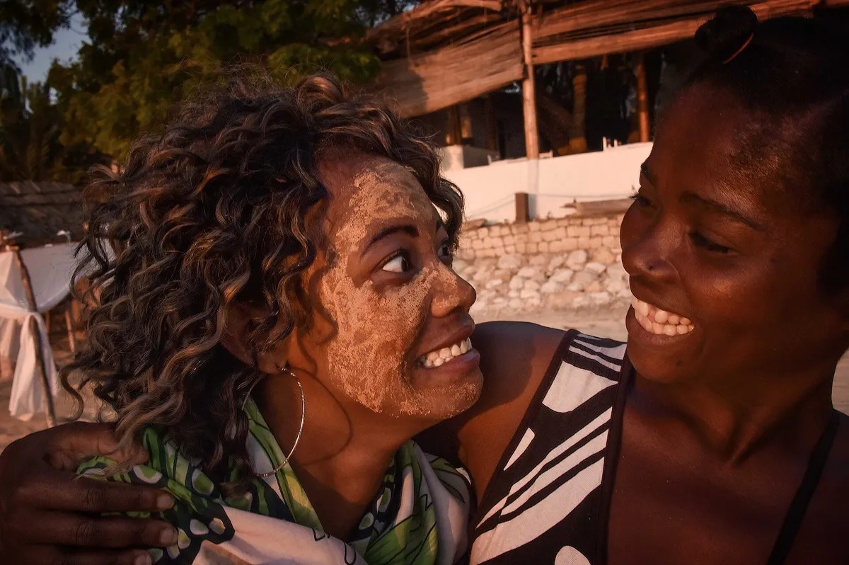 Duas amigas sorrindo abraçadas na praia ao entardecer, uma delas com areia no rosto depois de brincar