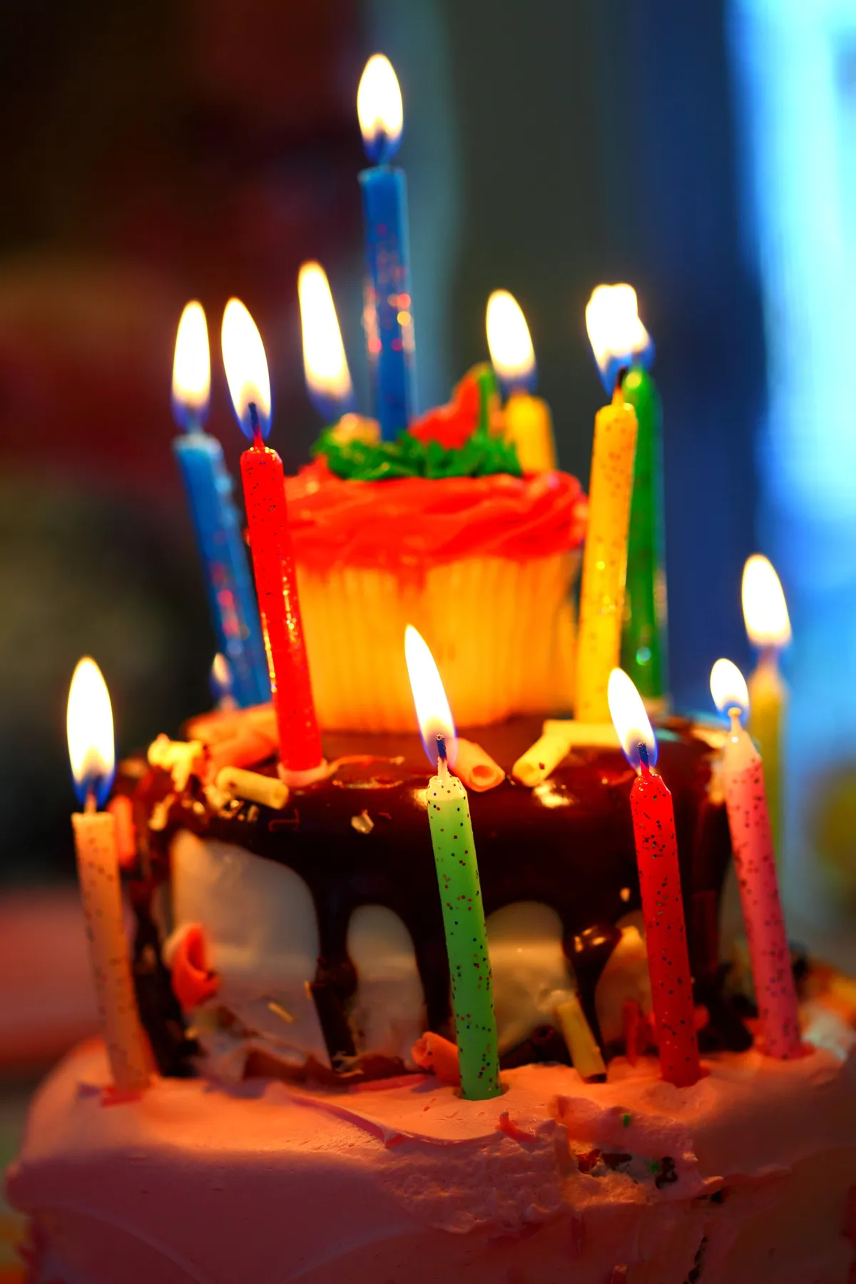 Bolo de aniversário com velas coloridas acesas em close, com um cupcake ao fundo