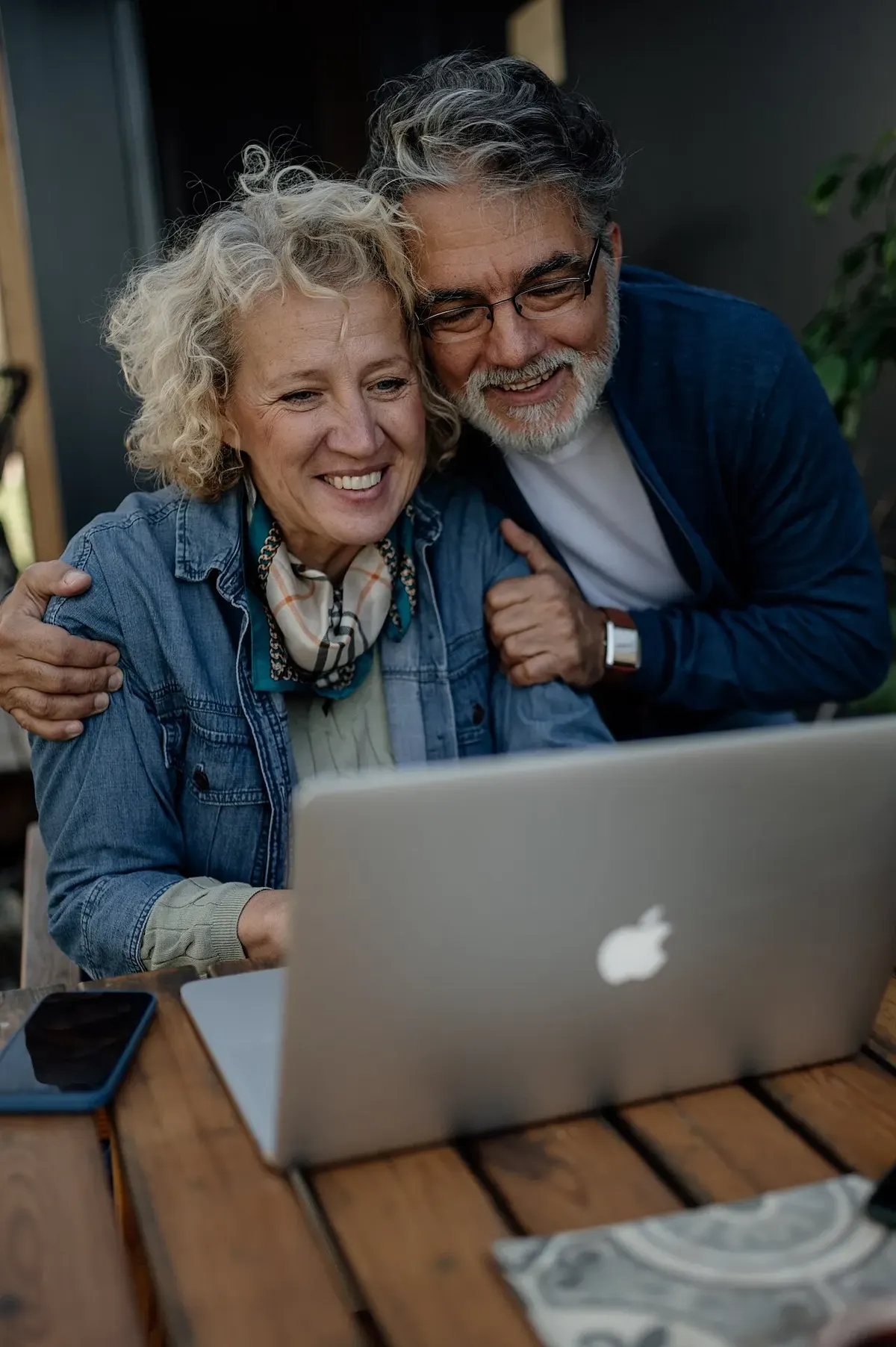 Casal de idosos sorrindo enquanto usa um laptop em uma mesa de madeira, consultando serviços pela internet