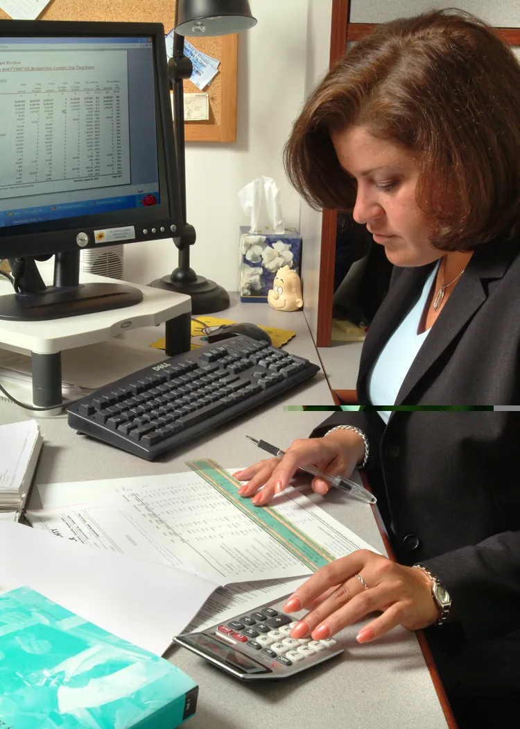 Profissional conferindo documentos impressos sobre a mesa, com régua e calculadora ao lado de um computador
