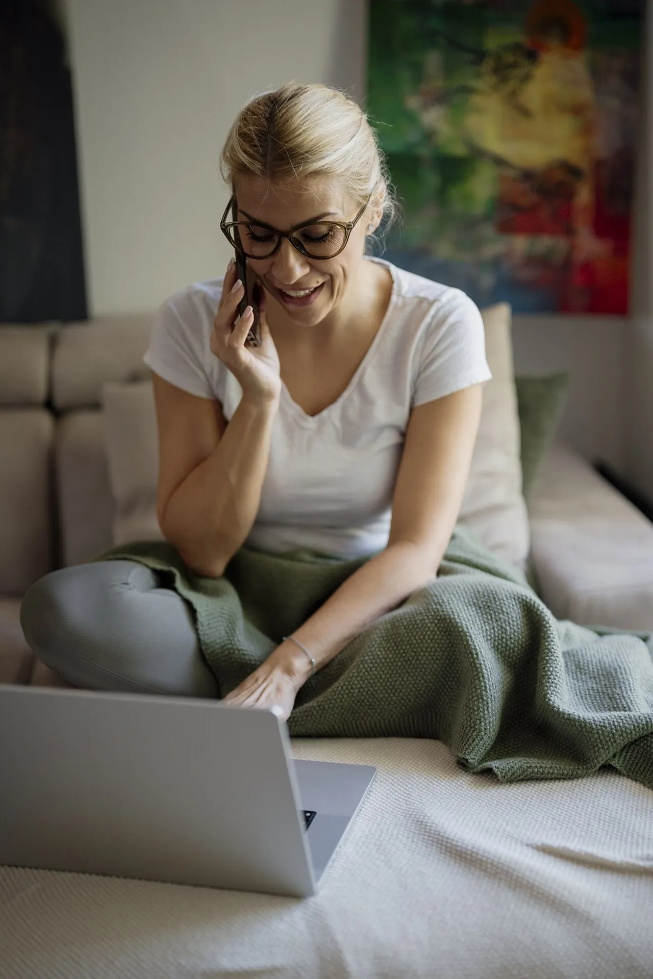 Una mujer sentada en un sillón consulta su laptop mientras sostiene su teléfono