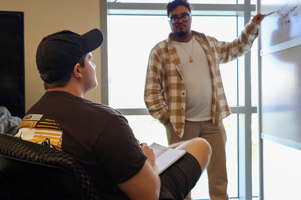 Estudiante tomando notas en un cuaderno mientras un instructor explica un concepto en un pizarrón