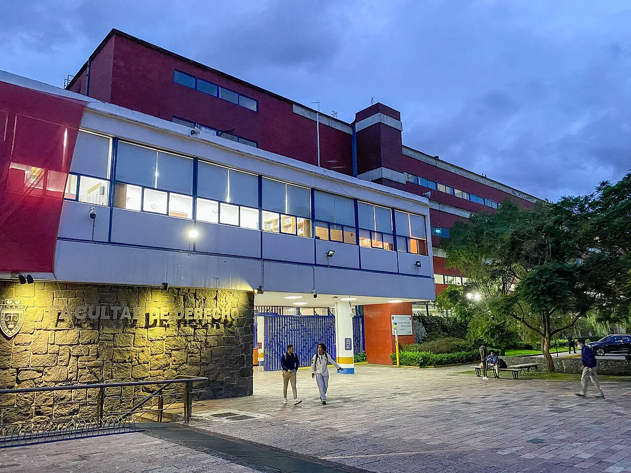 Fachada de la Facultad de Derecho de la UNAM al anochecer con estudiantes en la explanada