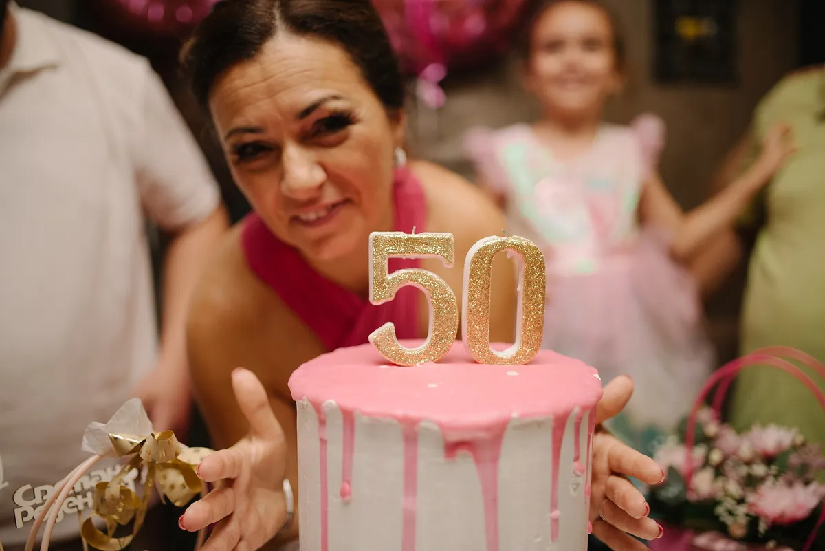 Mulher sorrindo atrás de um bolo rosa com velas douradas de 50 anos, em festa de aniversário com a família