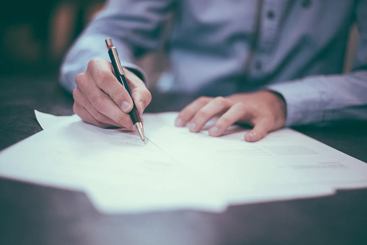 Una mano firmando un documento sobre una mesa, junto a una pluma