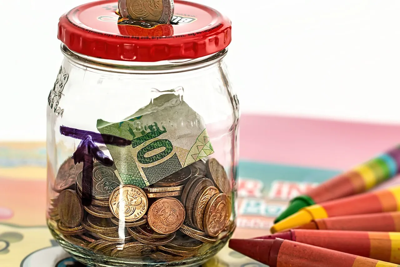 Un frasco de vidrio con monedas y un billete, junto a crayones de colores