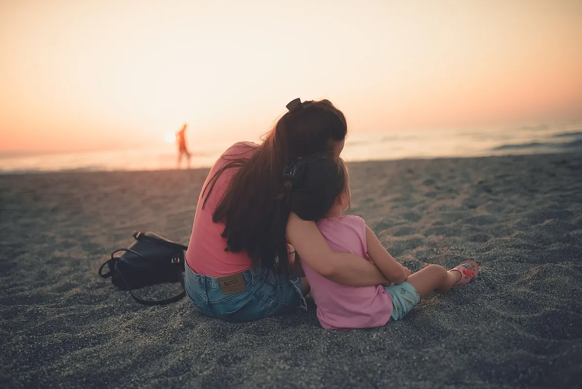 Mãe e filha pequena abraçadas sentadas na areia da praia, de costas, olhando o pôr do sol no mar