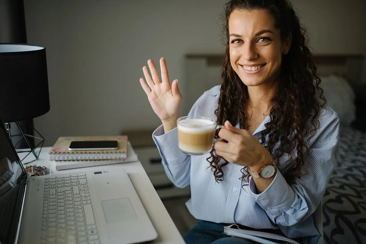 Una mujer trabaja en su laptop en una mesa con una taza de café, saludando a la cámara
