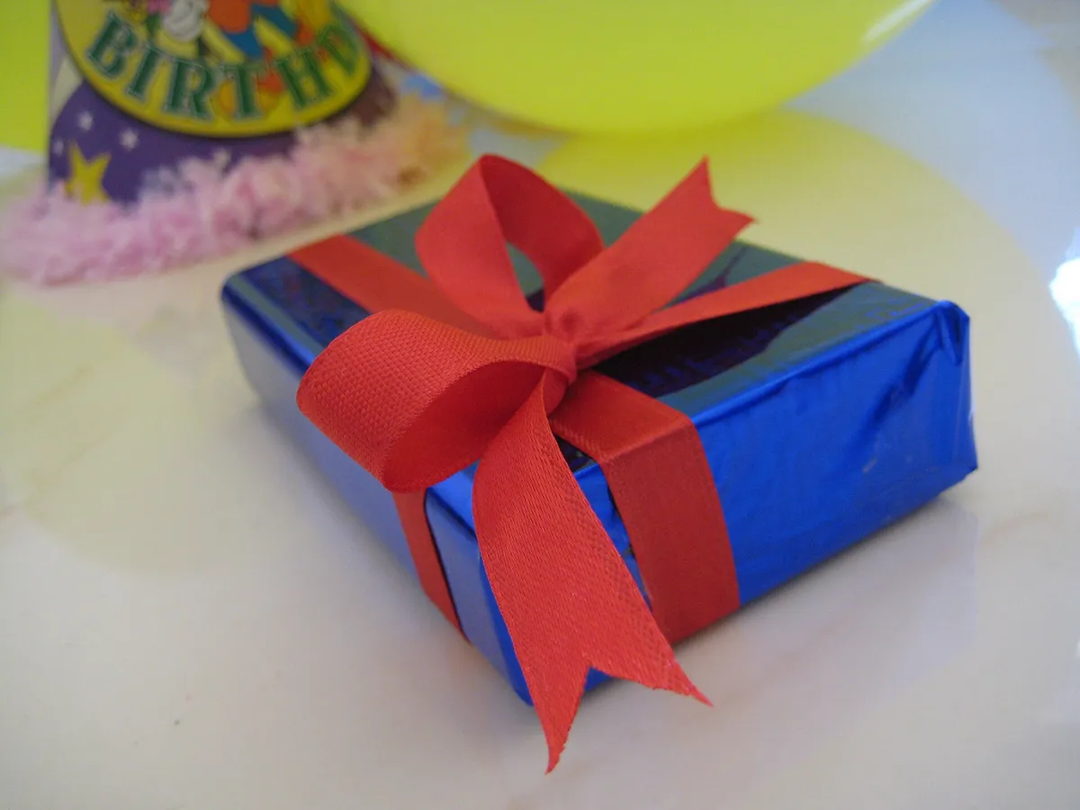 Caixa de presente azul com laço vermelho sobre a mesa, ao lado de balão e chapéu de festa