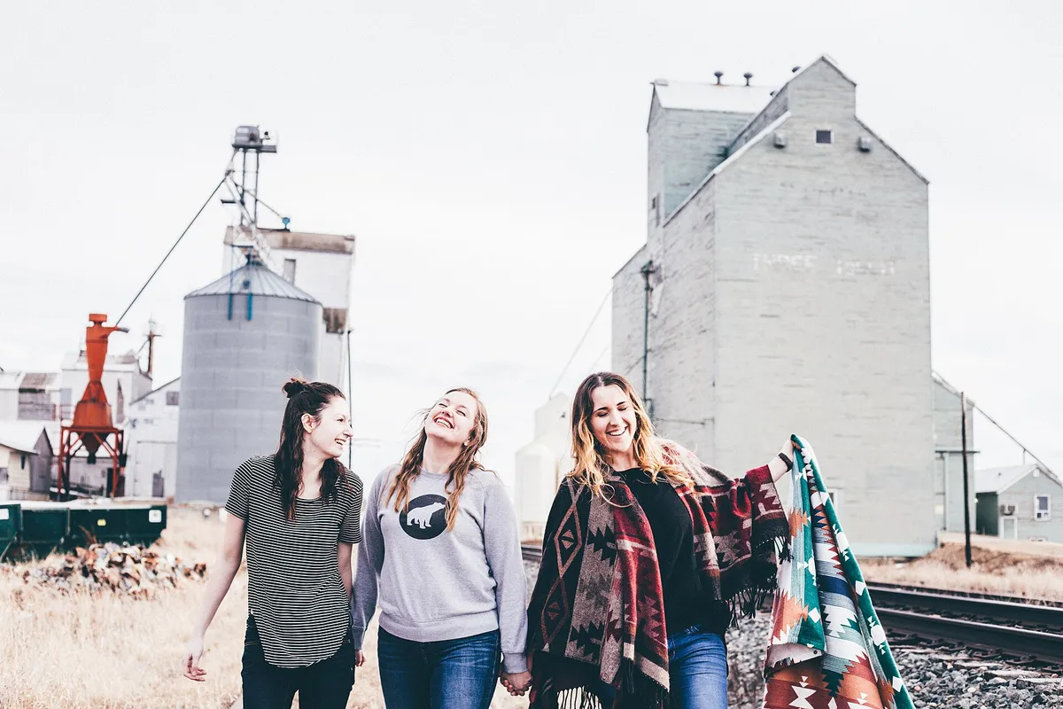 Três amigas jovens caminhando e rindo ao lado de trilhos de trem, em cidade do interior
